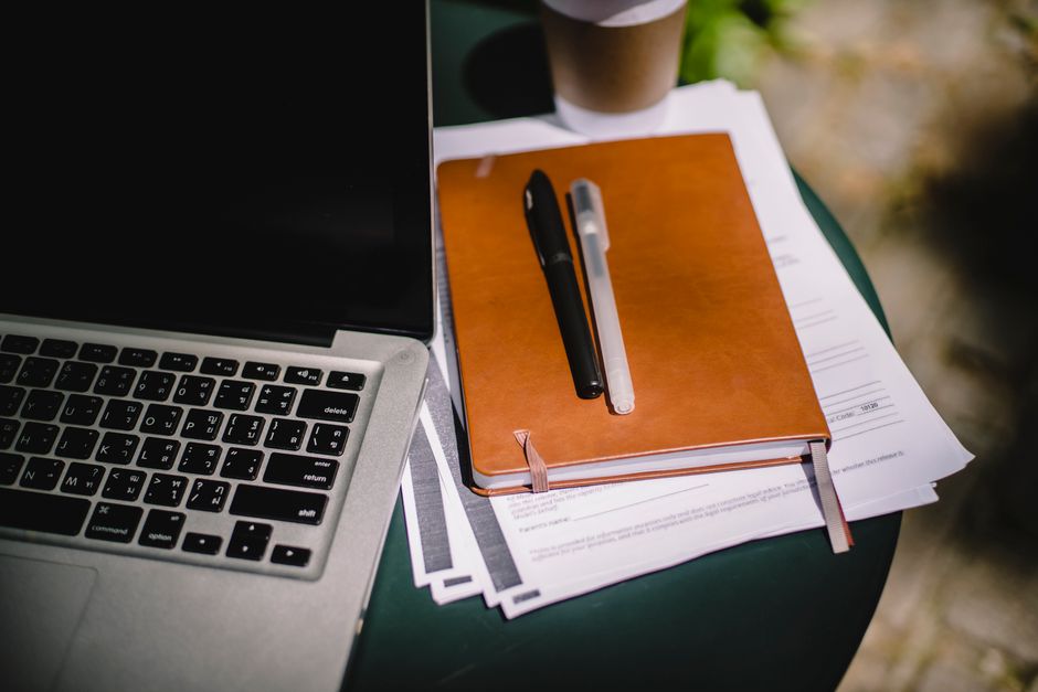 A leather notebook, pens and paperwork beside a laptop